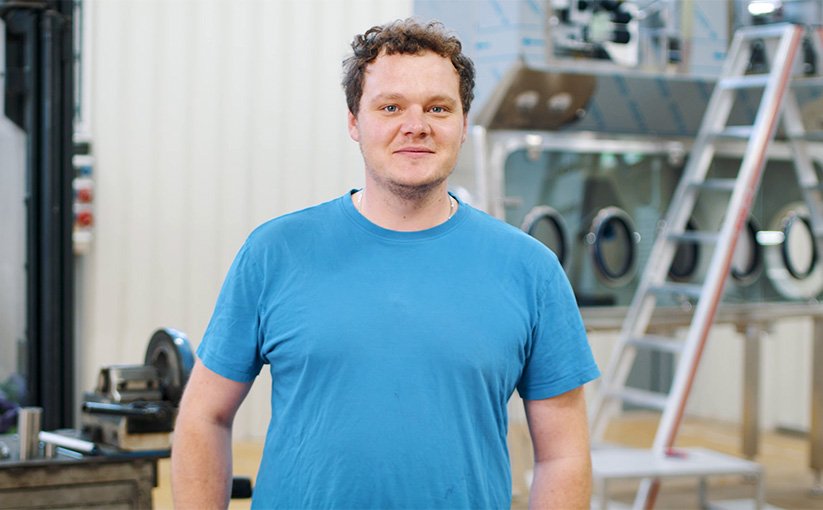Mann in blauem T-Shirt spricht in einer Werkshalle, im Hintergrund sind Leitern und Maschinen sichtbar.